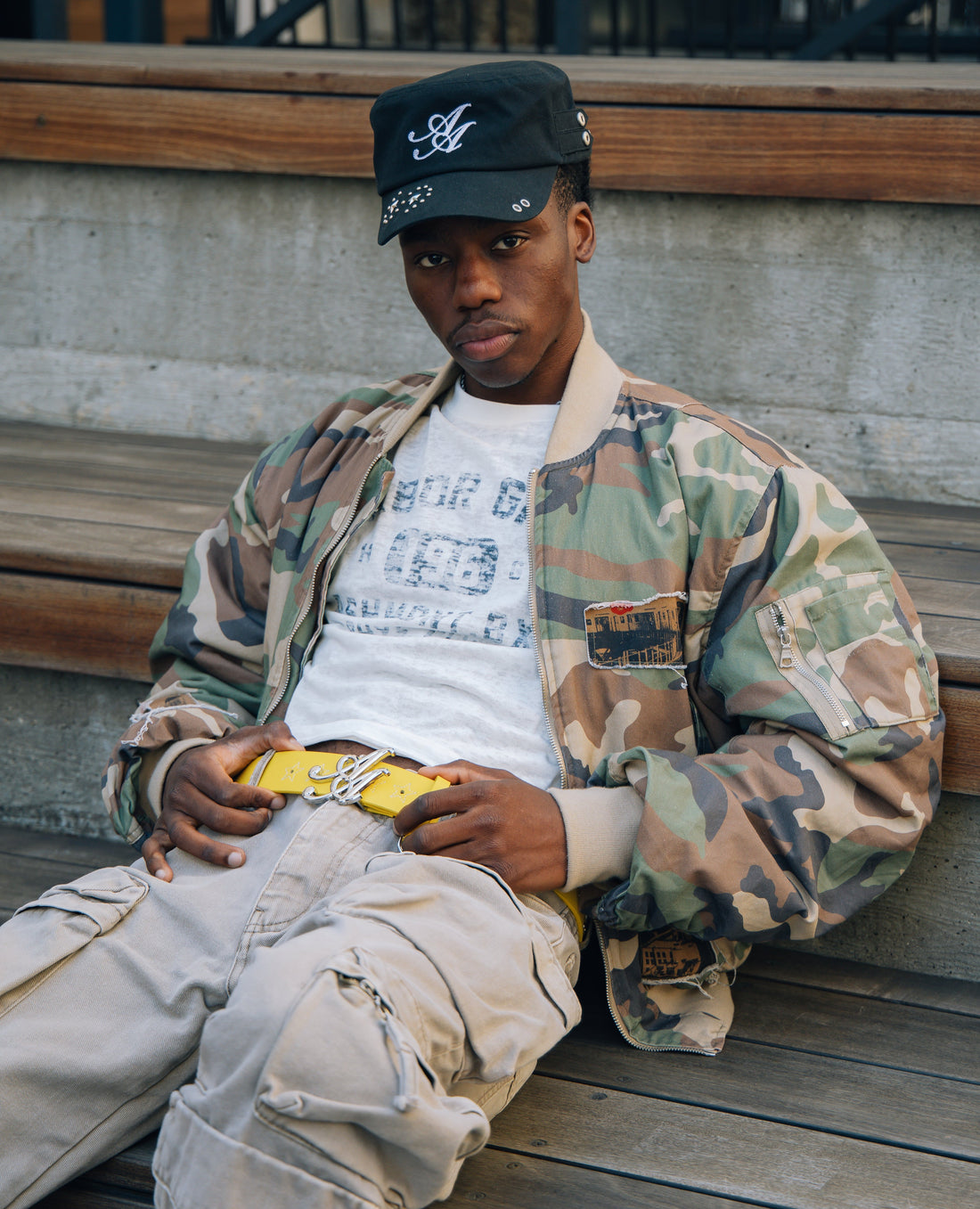 Model wearing the Ad Astra black cadet hat with a camo jacket and yellow Vellum belt